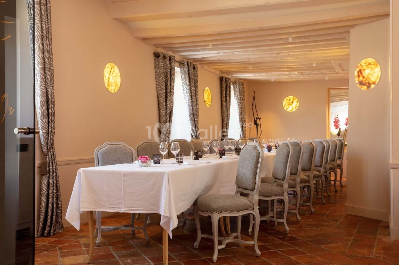 Salle à manger avec une longue table dressée, chaises élégantes, rideaux aux fenêtres et éclairage chaleureux.