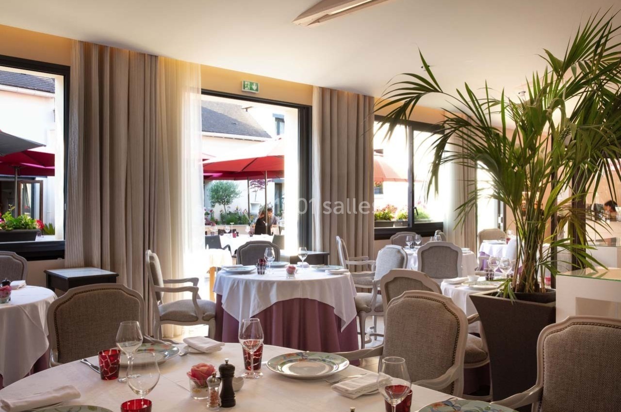 Salle de restaurant lumineuse avec tables dressées, grandes fenêtres ouvertes sur une terrasse avec parasols rouges.