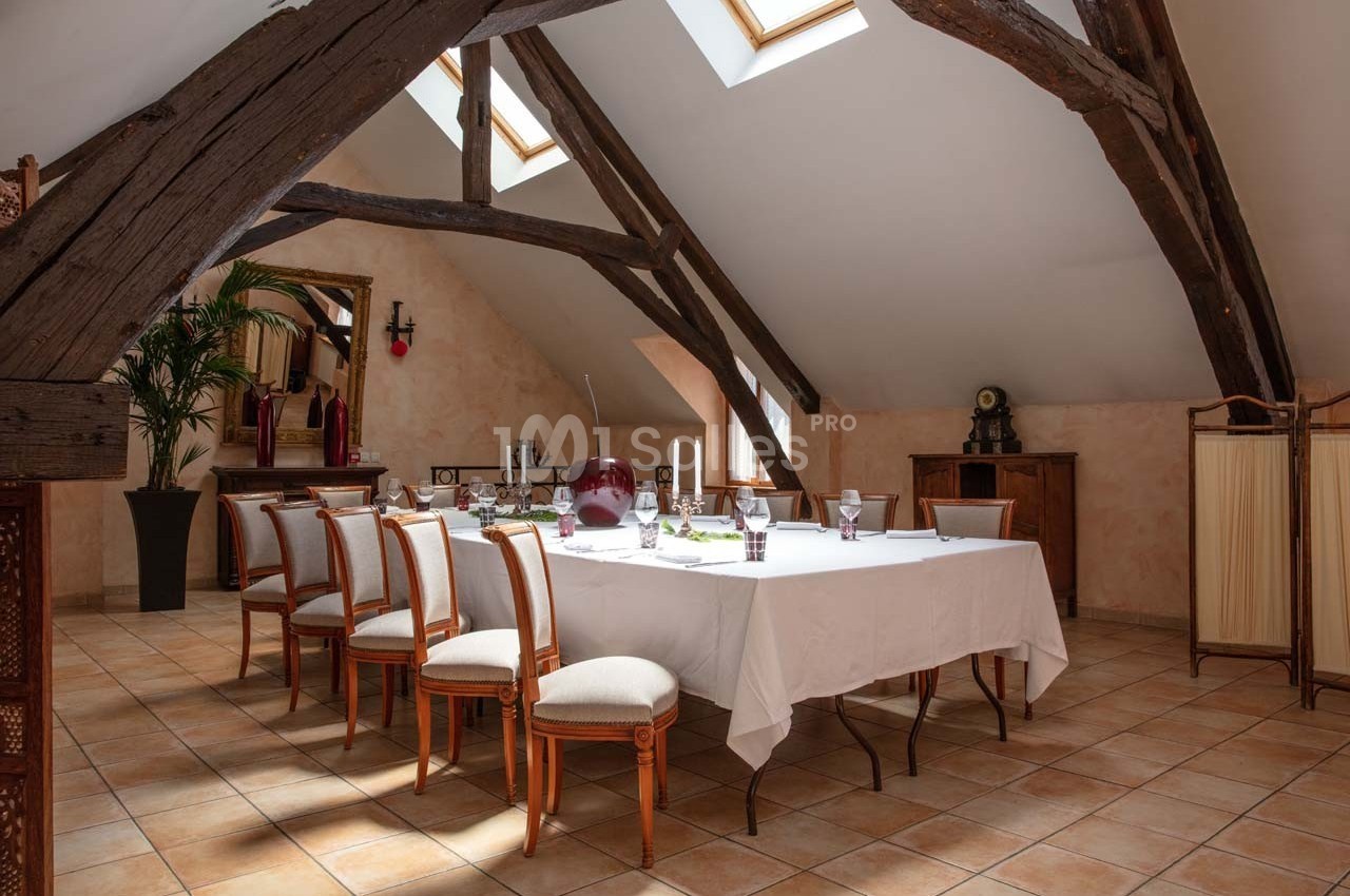 Salle lumineuse sous combles avec poutres apparentes, table dressée pour un repas, chaises en bois et décoration élégante.