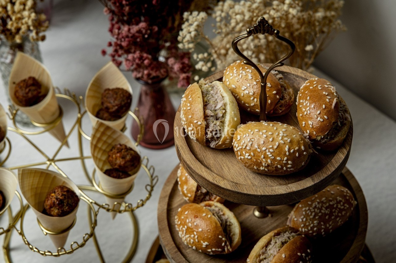 Plateau en bois avec des mini-burgers au sésame et des cônes en bois garnis de bouchées, décoré de fleurs séchées.