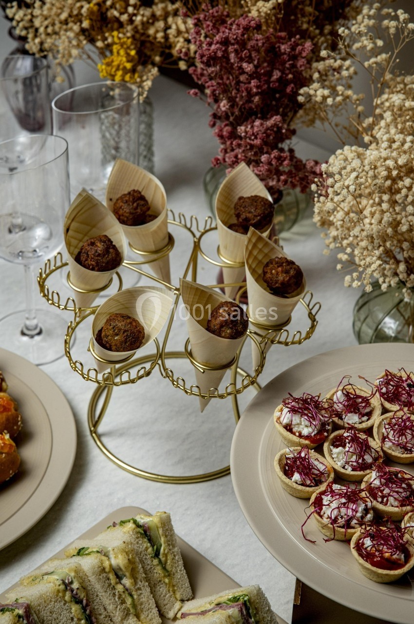 Présentoir doré avec amuse-bouches, entouré de sandwichs, tartelettes garnies et décorations florales en arrière-plan.
