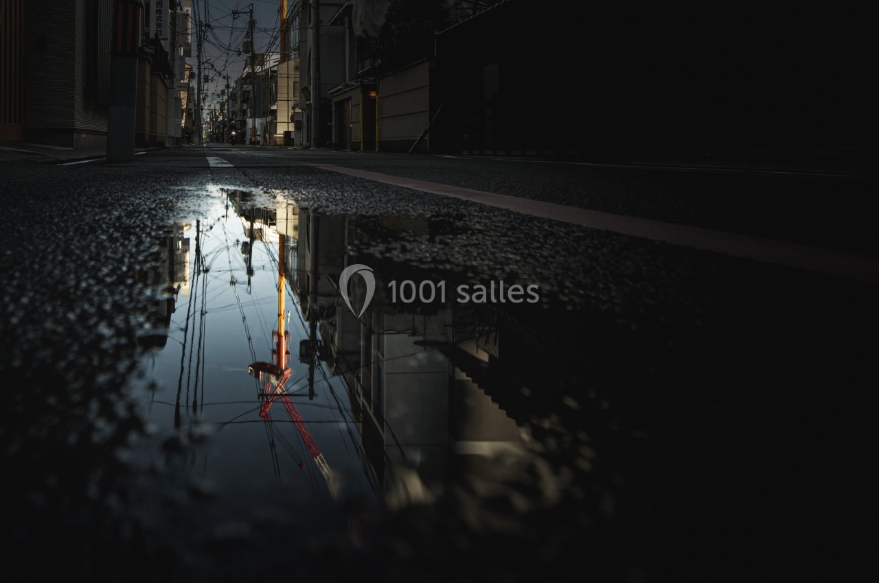 Reflet d'une rue urbaine et d'une structure métallique rouge dans une flaque d'eau au crépuscule.