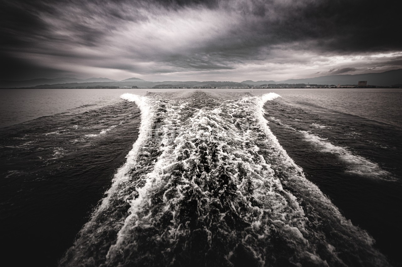 Sillage d'un bateau sur une étendue d'eau sombre sous un ciel nuageux, avec des montagnes en arrière-plan.