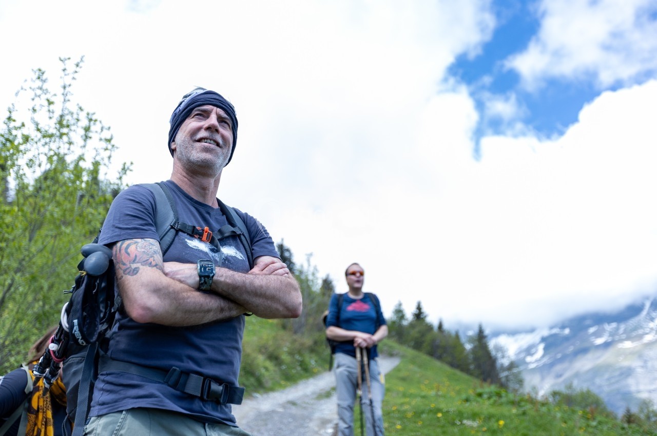 Un homme souriant avec un sac à dos sur un sentier de montagne, accompagné d'une autre personne en arrière-plan.