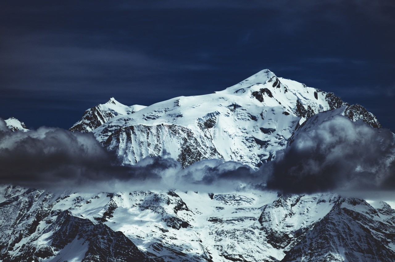 Montagne enneigée sous un ciel sombre, partiellement enveloppée de nuages bas.