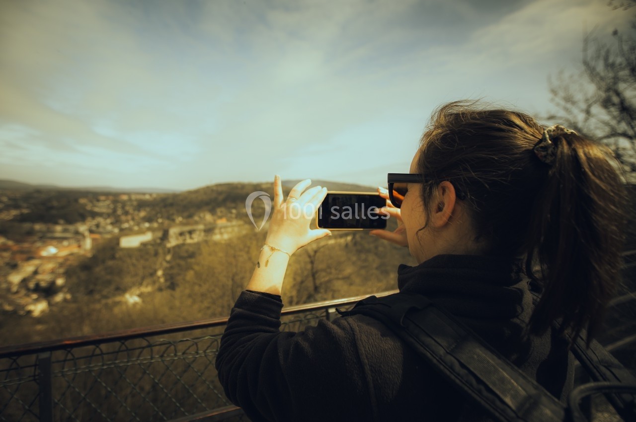 Une femme photographie un paysage vallonné depuis un point de vue, appuyée contre une barrière métallique.
