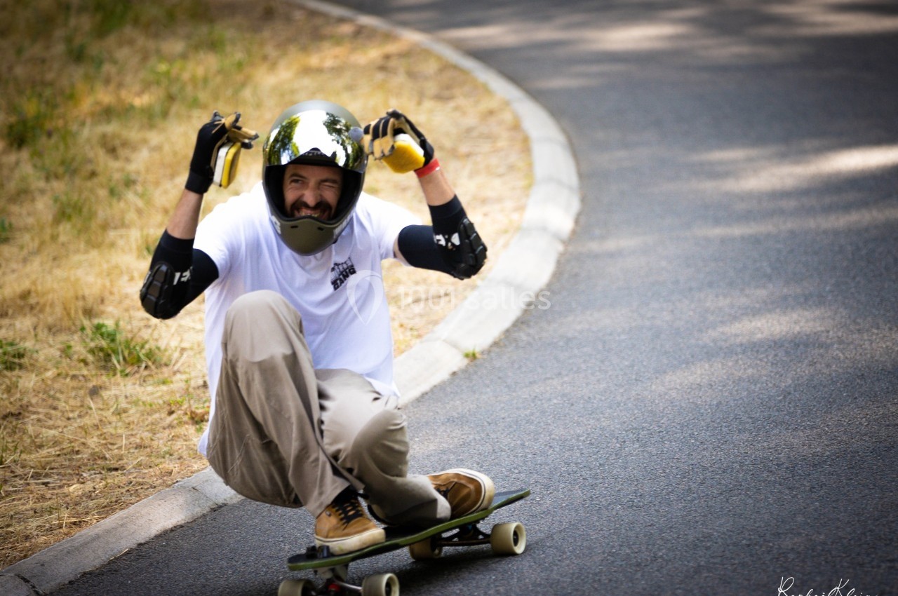 Un skateur en descente sur une route, portant un casque et des protections, fait un geste amusant avec les mains.