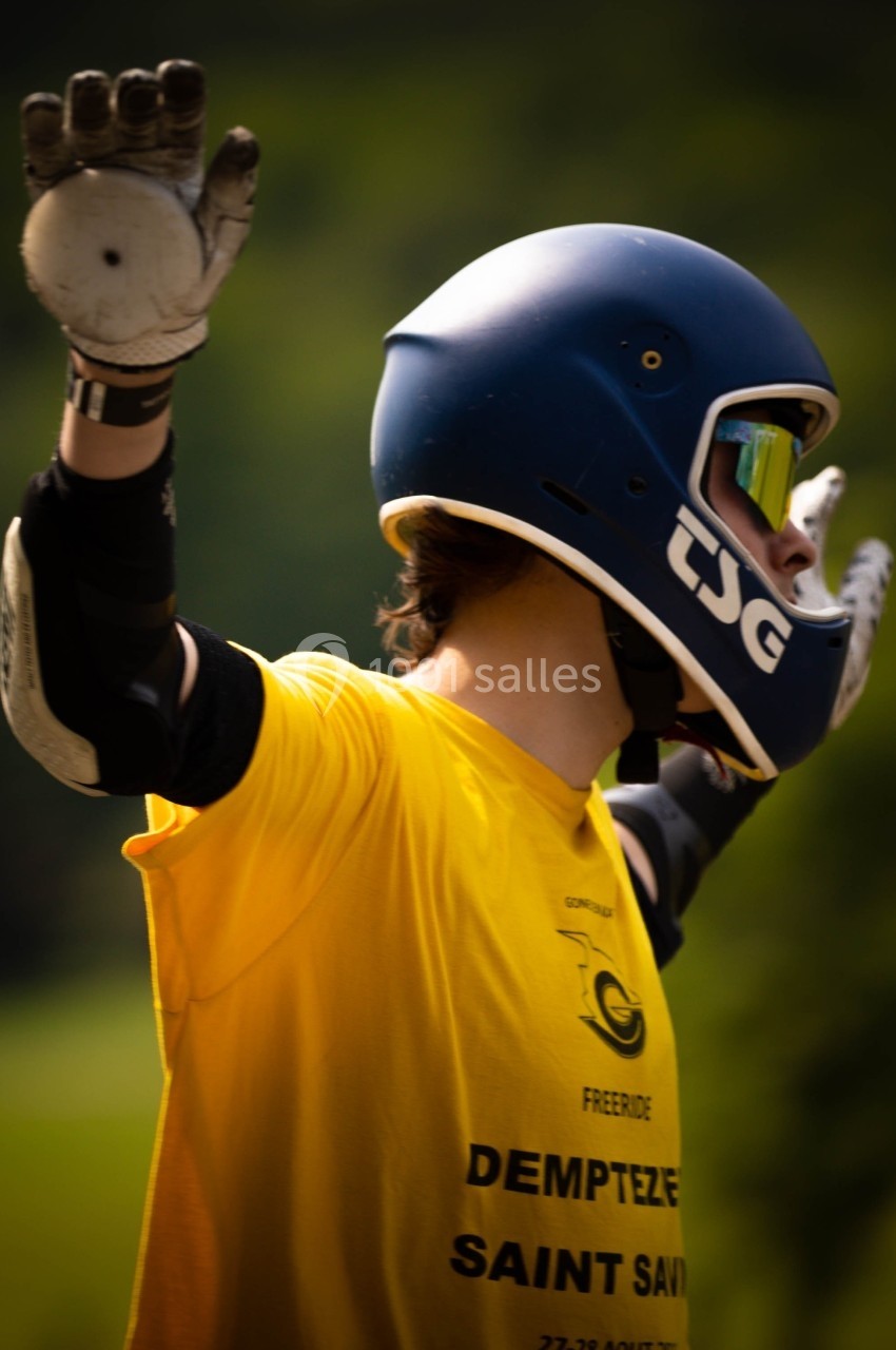 Personne portant un casque et des gants, les bras levés, vêtue d'un t-shirt jaune, en extérieur verdoyant.