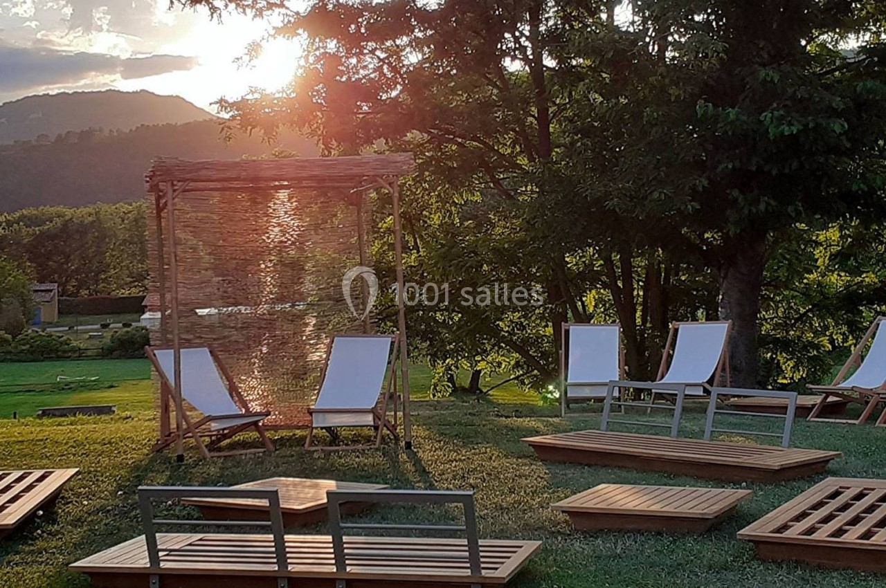 Chaises longues et tables en bois disposées sur une pelouse, avec un soleil couchant et des arbres en arrière-plan.