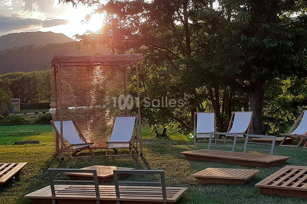Chaises longues et tables en bois disposées sur une pelouse, avec un soleil couchant et des arbres en arrière-plan.