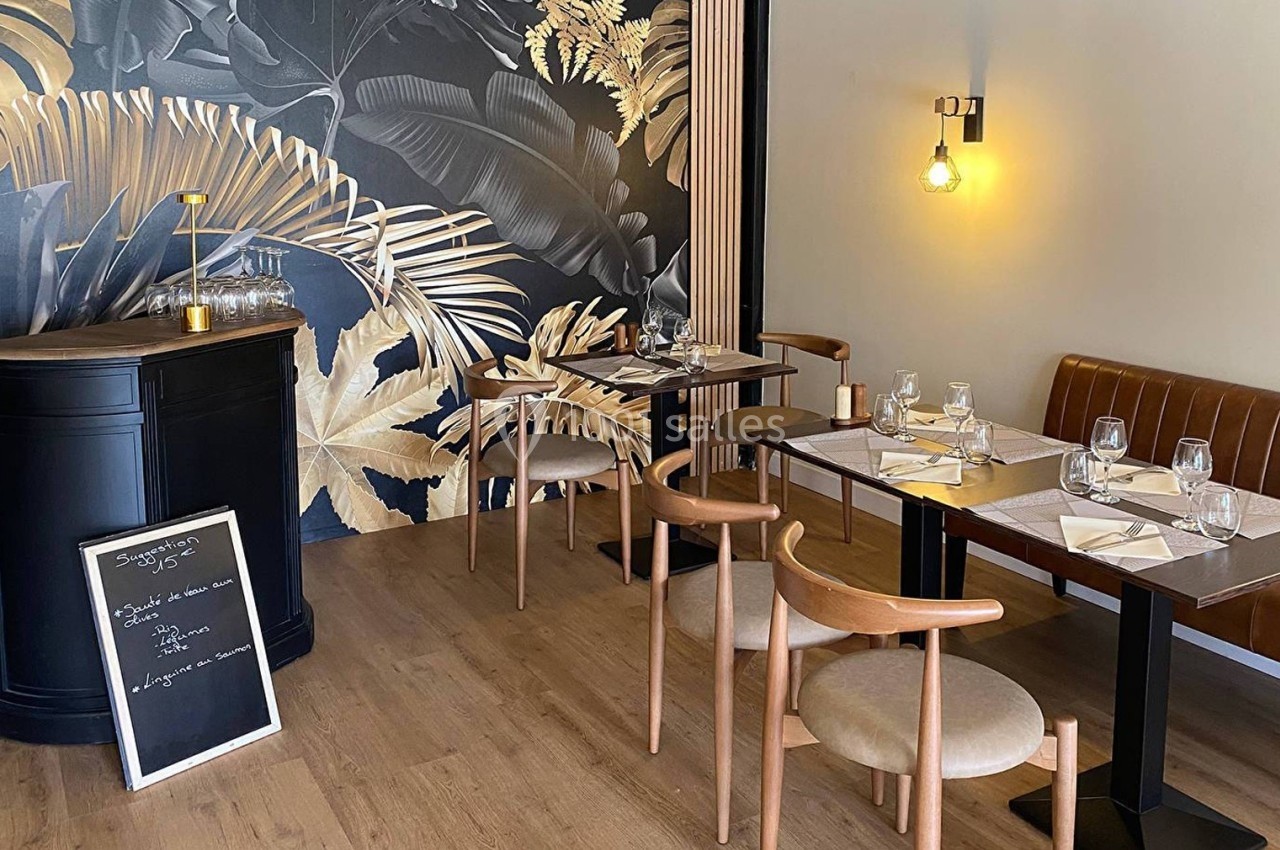 Salle de restaurant avec tables dressées, chaises en bois, mur décoré de motifs végétaux et éclairage chaleureux.