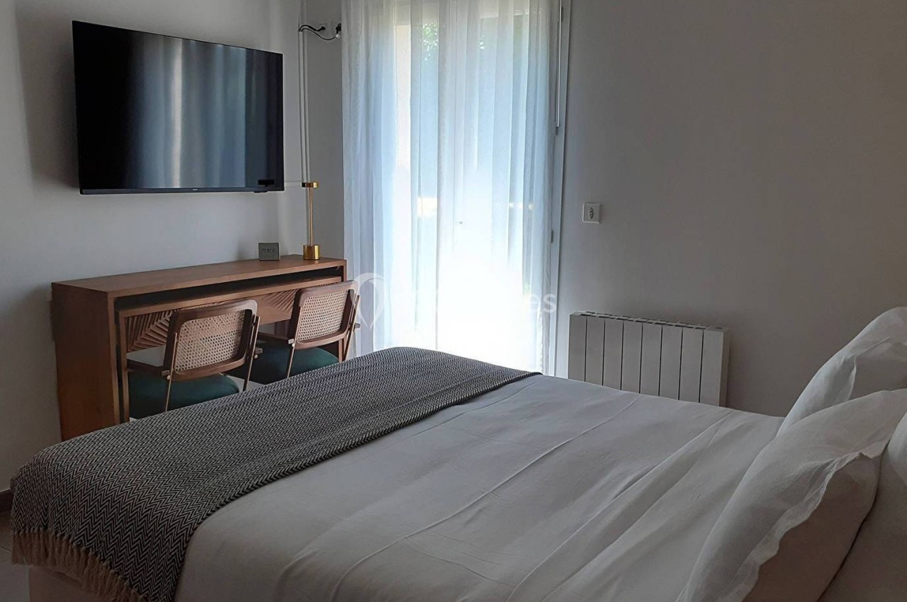 Chambre lumineuse avec lit double, télévision murale, bureau en bois, deux chaises et rideaux blancs filtrant la lumière.