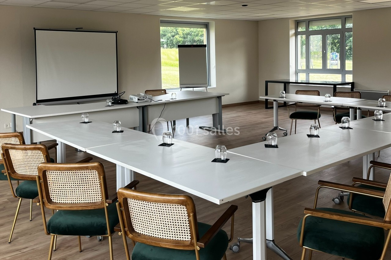 Salle de réunion lumineuse avec tables disposées en U, chaises en bois et écran de projection à l'avant.