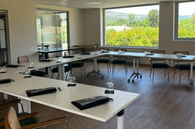 Chambre lumineuse avec lit double, télévision murale, bureau en bois, deux chaises et rideaux blancs filtrant la lumière.