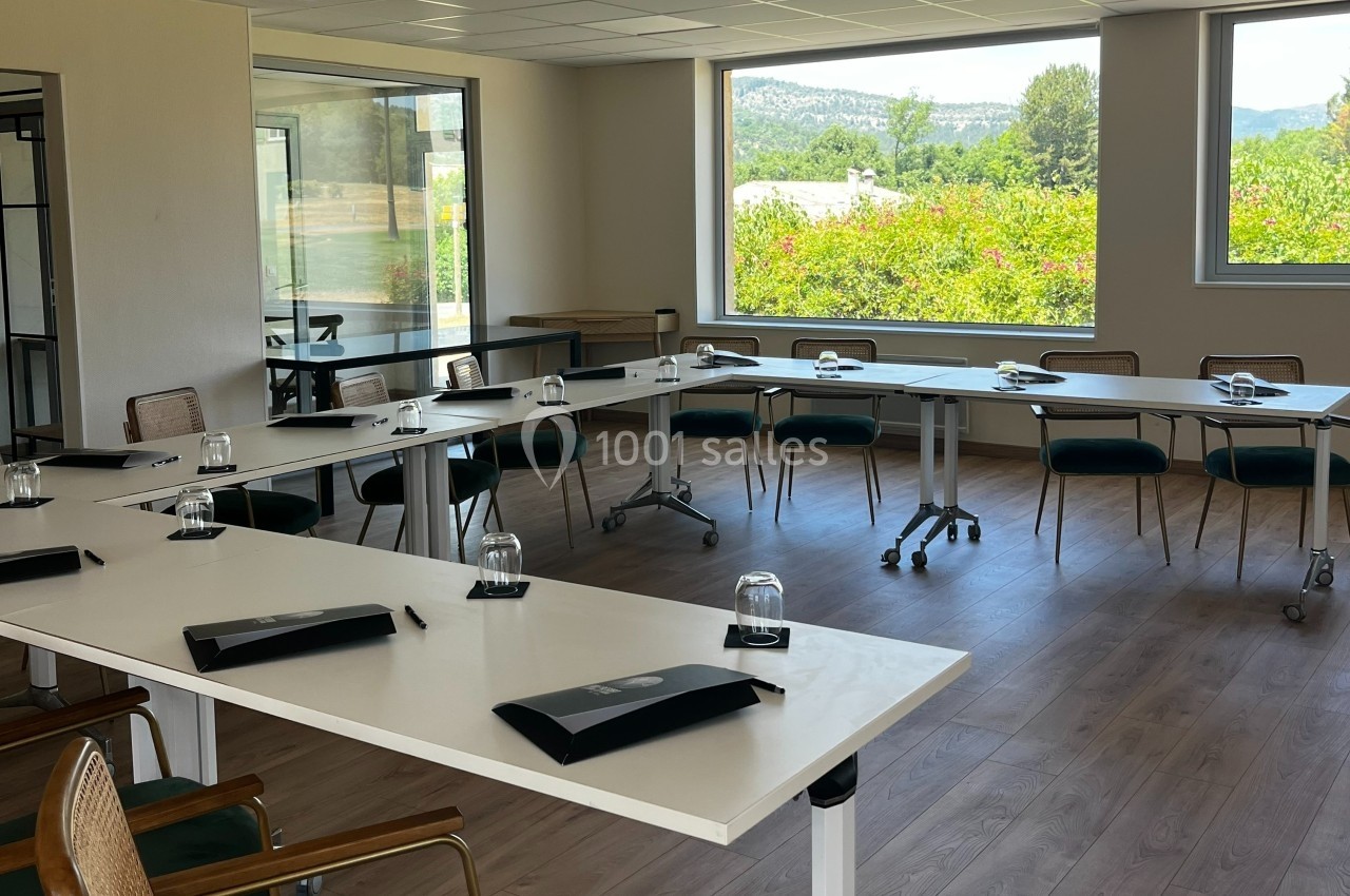 Salle de réunion lumineuse avec tables disposées en U, chaises, blocs-notes et verres, vue sur un paysage verdoyant.