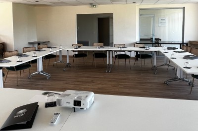 Chambre lumineuse avec lit double, télévision murale, bureau en bois, deux chaises et rideaux blancs filtrant la lumière.