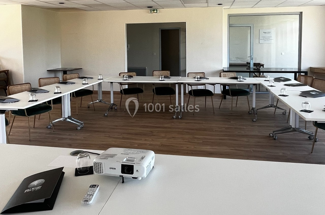 Salle de réunion lumineuse avec tables disposées en U, chaises, vidéoprojecteur et documents sur les tables.