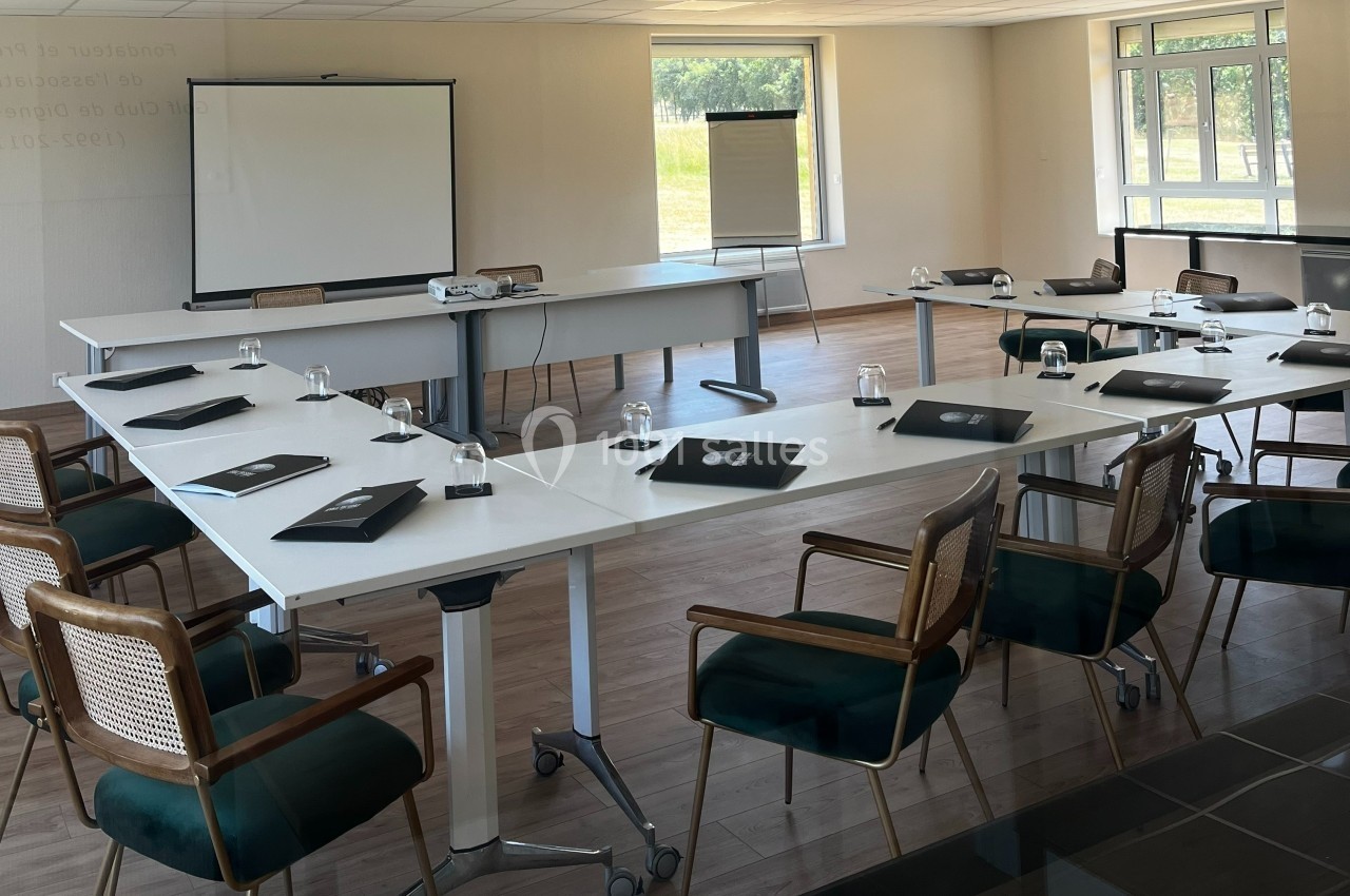 Salle de réunion lumineuse avec tables en U, chaises en bois, écran de projection et paperboard près des fenêtres.