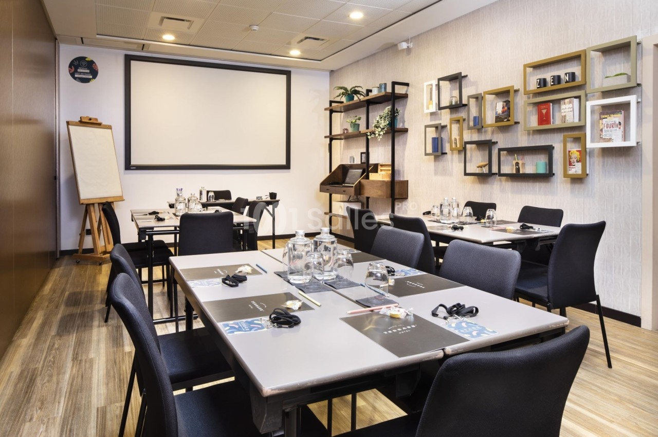 Salle de réunion équipée avec tables, chaises, écran de projection, tableau blanc et étagères décoratives.