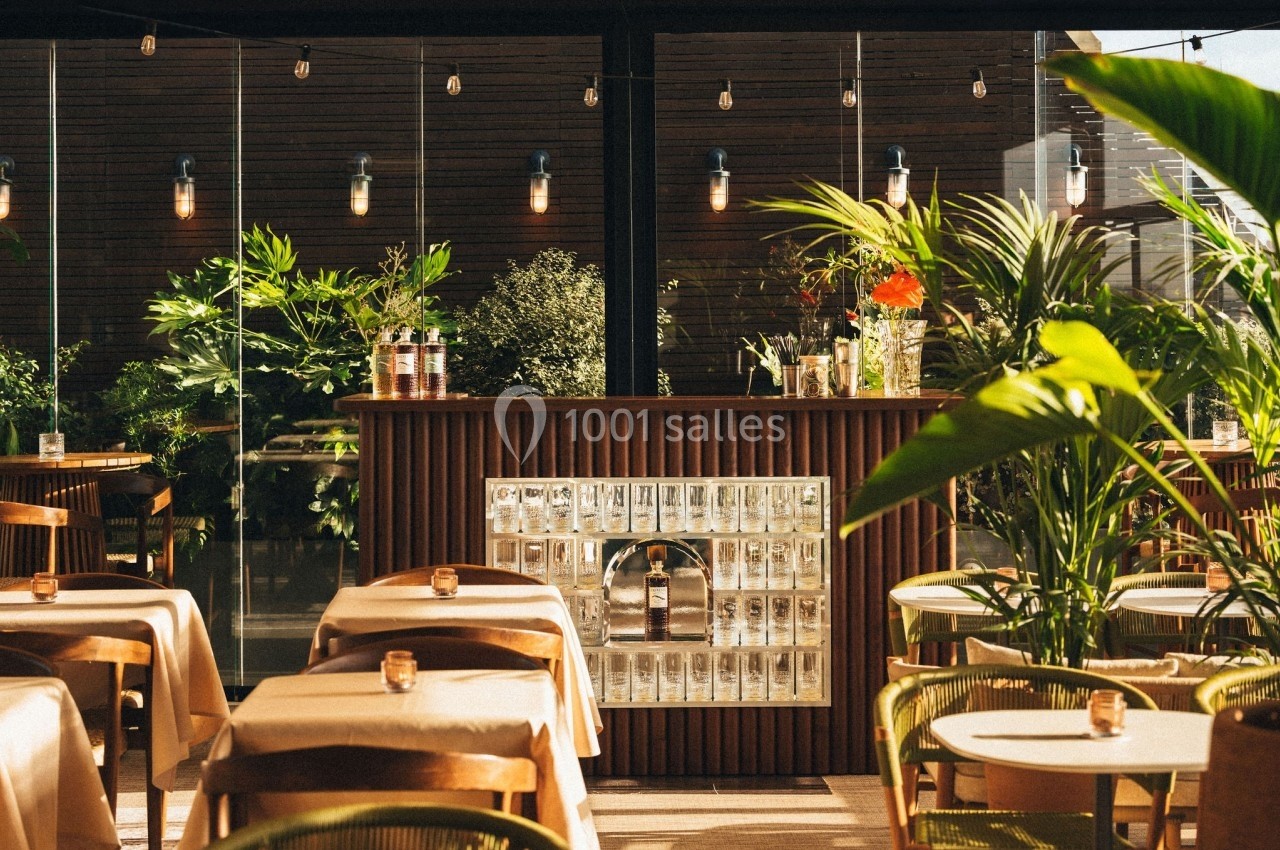 Terrasse lumineuse avec tables dressées, bar en bois central, plantes vertes et éclairage suspendu.