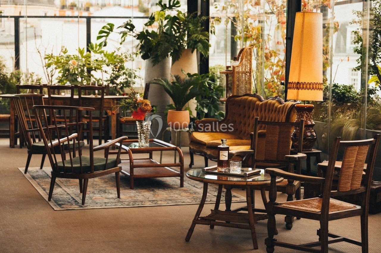Salon lumineux avec mobilier vintage, plantes vertes, tapis et grande lampe, donnant sur un espace extérieur.