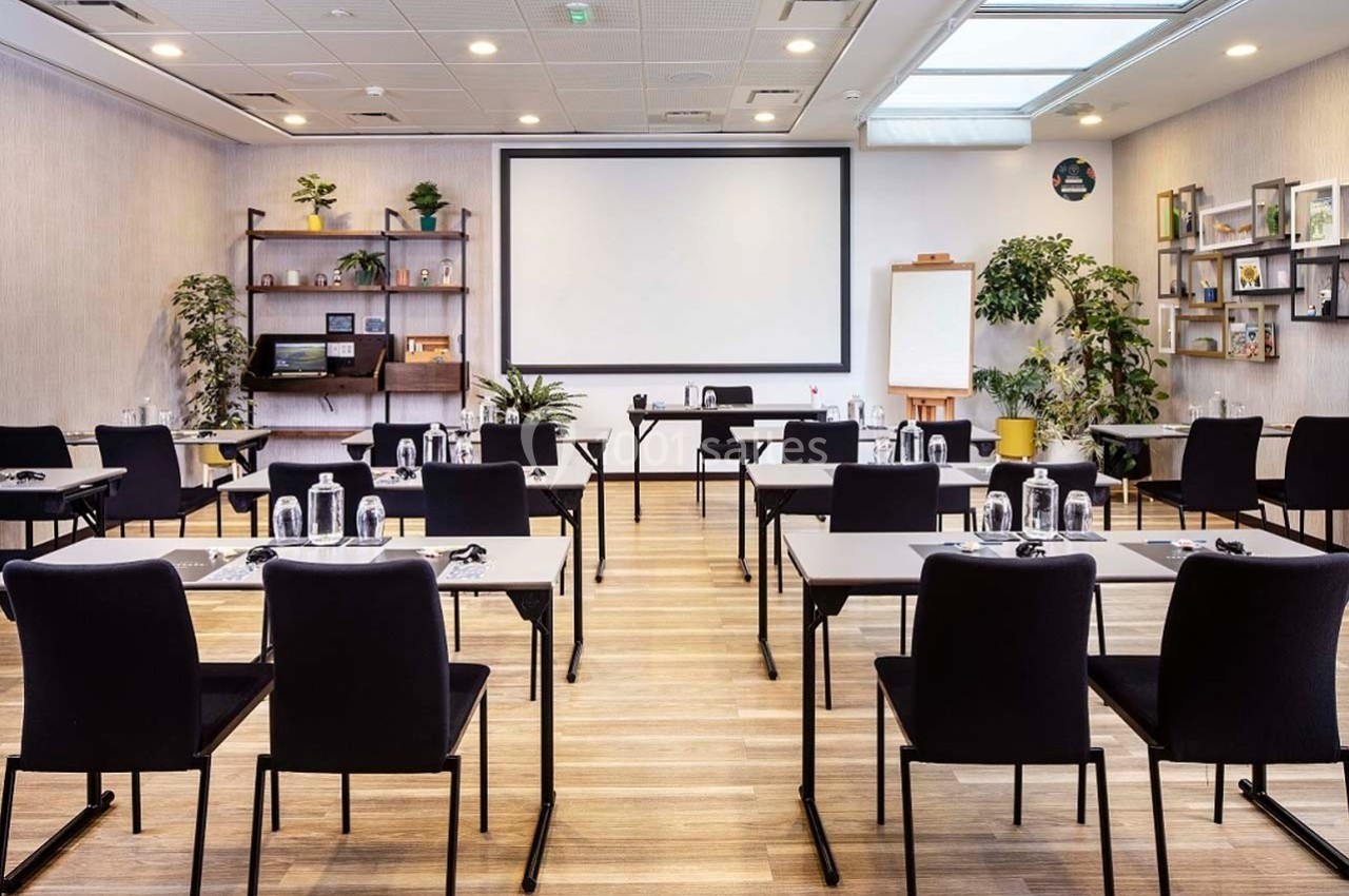 Salle de réunion lumineuse avec des tables et chaises alignées, un écran de projection et des plantes décoratives.