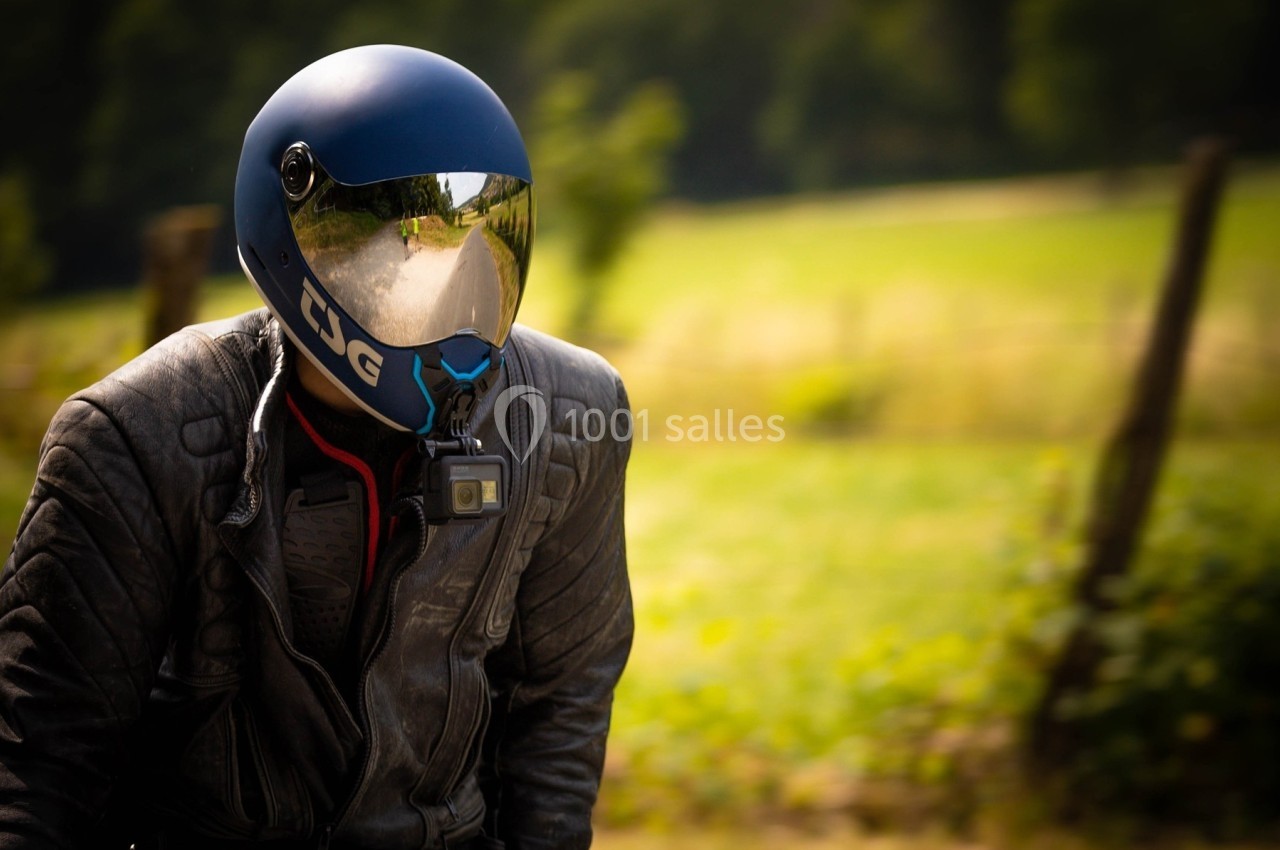 Motard portant un casque bleu avec visière réfléchissante, vêtu d'une veste en cuir, dans un paysage champêtre.
