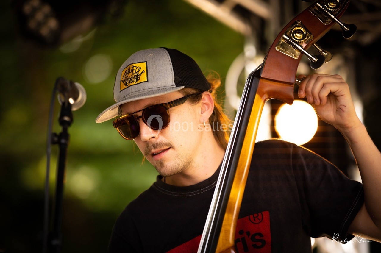 Un musicien joue de la contrebasse sur une scène en extérieur, portant des lunettes de soleil et une casquette.