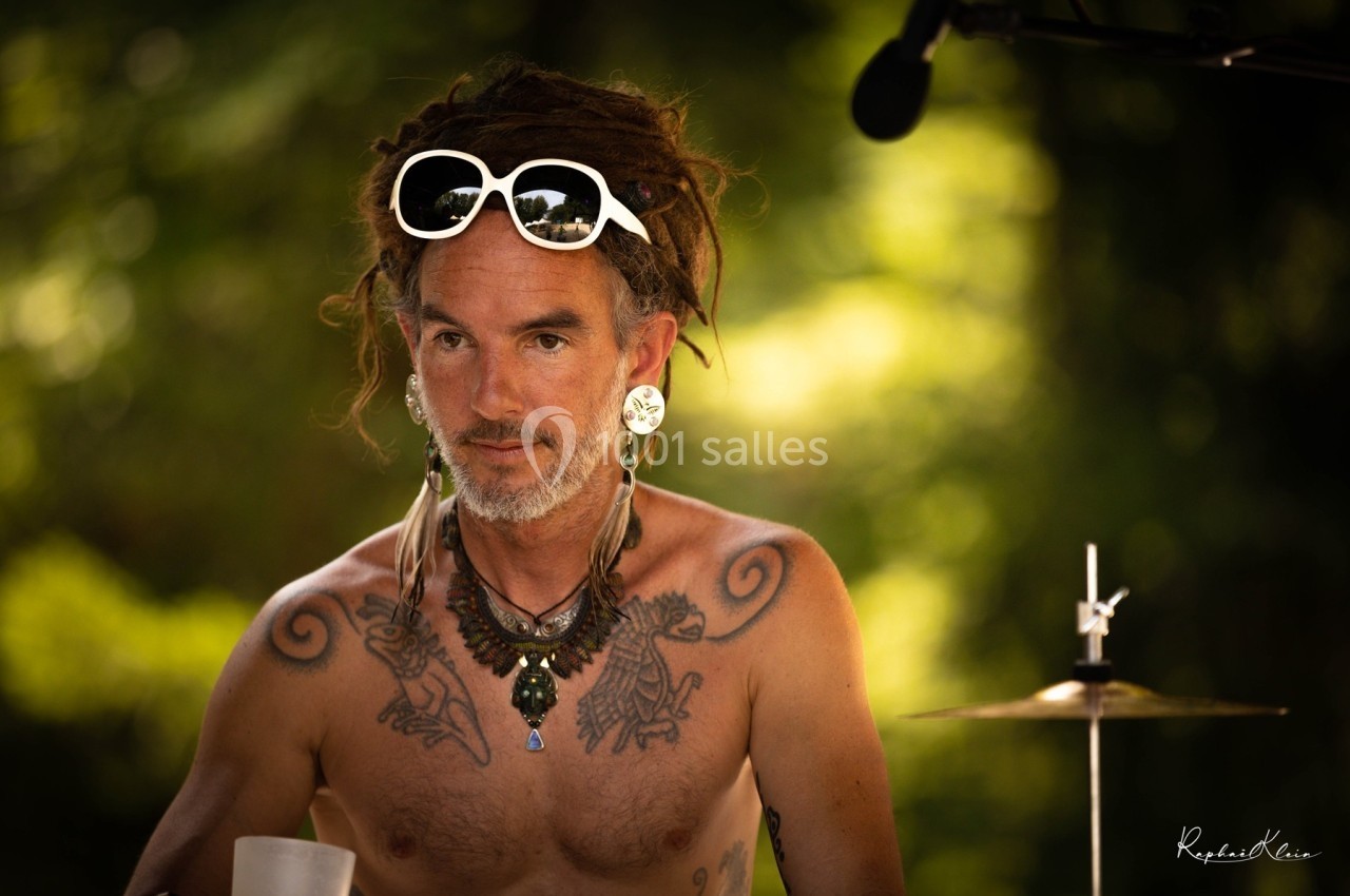 Homme torse nu avec des dreadlocks, lunettes blanches sur la tête, portant des bijoux, devant un fond naturel flou.