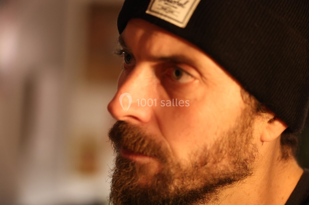Homme barbu portant un bonnet noir, regardant vers la gauche dans un environnement intérieur flou.