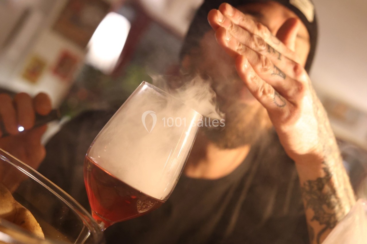 Un homme souffle de la fumée au-dessus d'un verre de vin rouge dans un environnement intérieur tamisé.