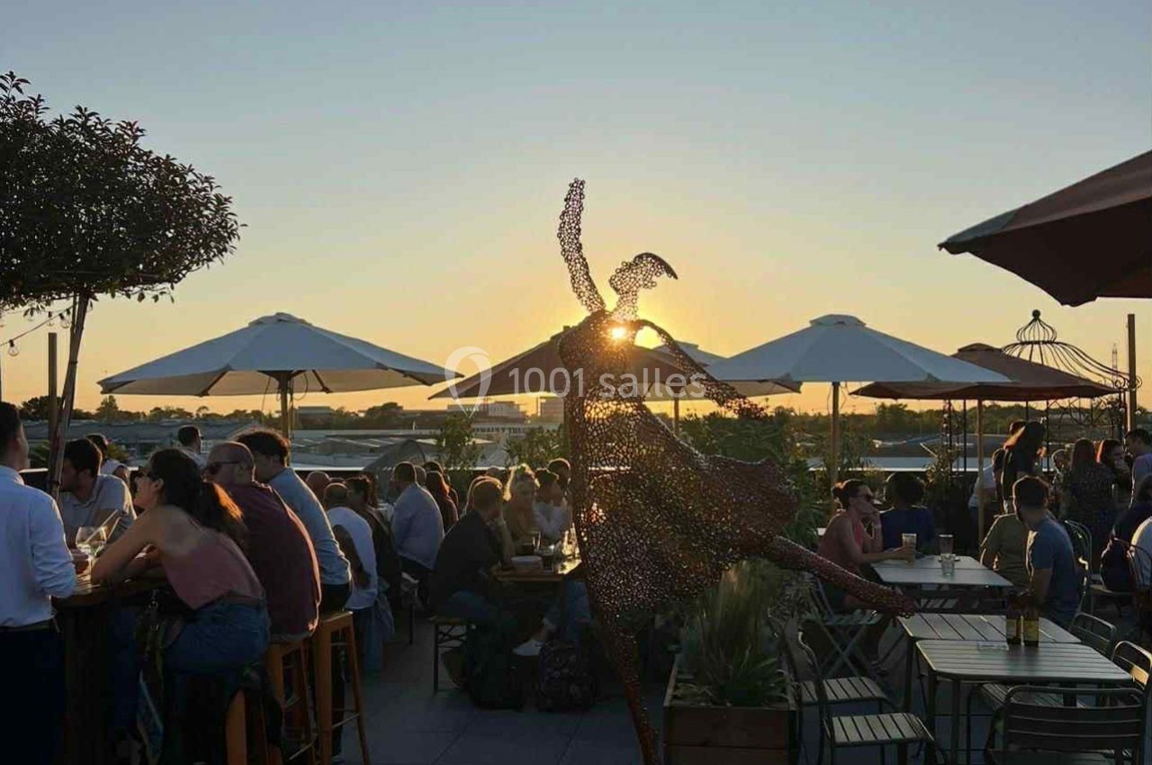 Statue d'une danseuse en métal sur une terrasse animée au coucher du soleil, entourée de parasols et de convives.