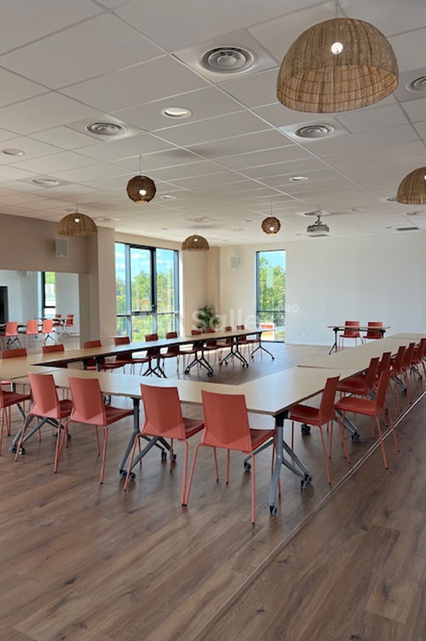 Salle lumineuse avec tables disposées en U, chaises rouges, parquet en bois et grandes fenêtres donnant sur l'extérieur.