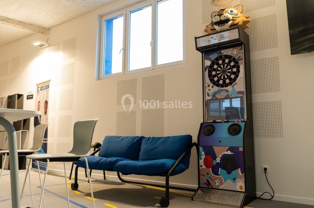 Un canapé bleu et une borne de jeu avec cible de fléchettes dans une salle lumineuse au design moderne.