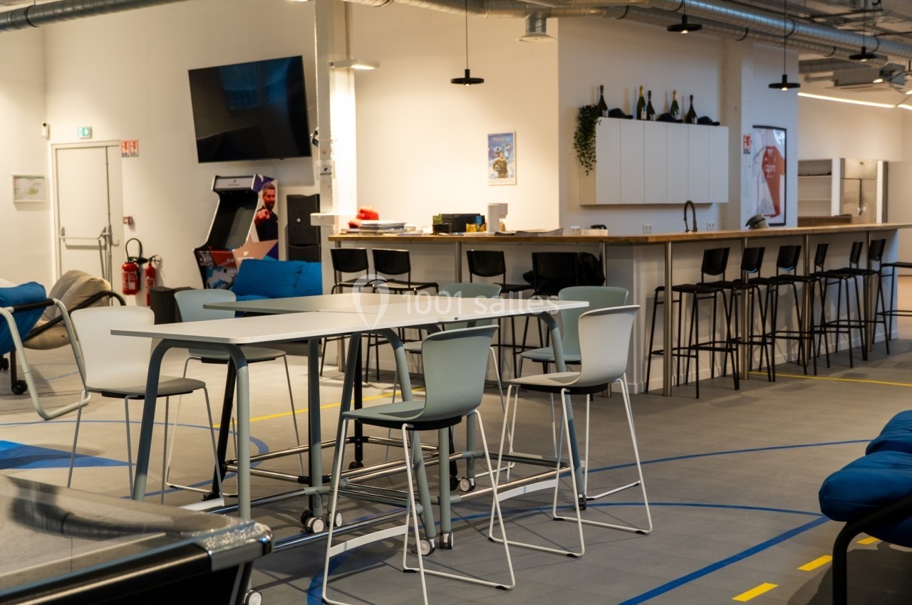 Espace de détente moderne avec tables hautes, chaises, canapé, et coin cuisine dans un environnement lumineux.
