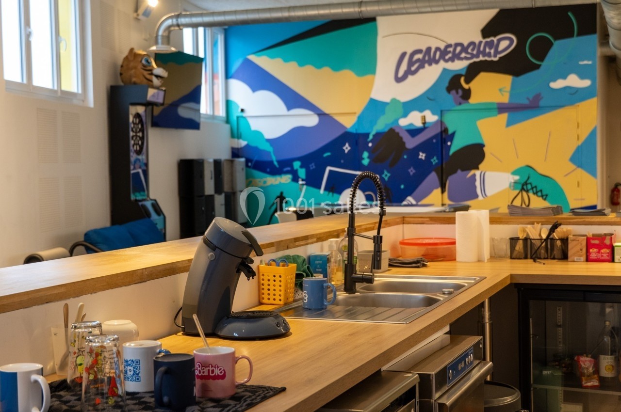 Cuisine partagée avec un comptoir en bois, évier, machine à café et mur décoré d'une fresque colorée.