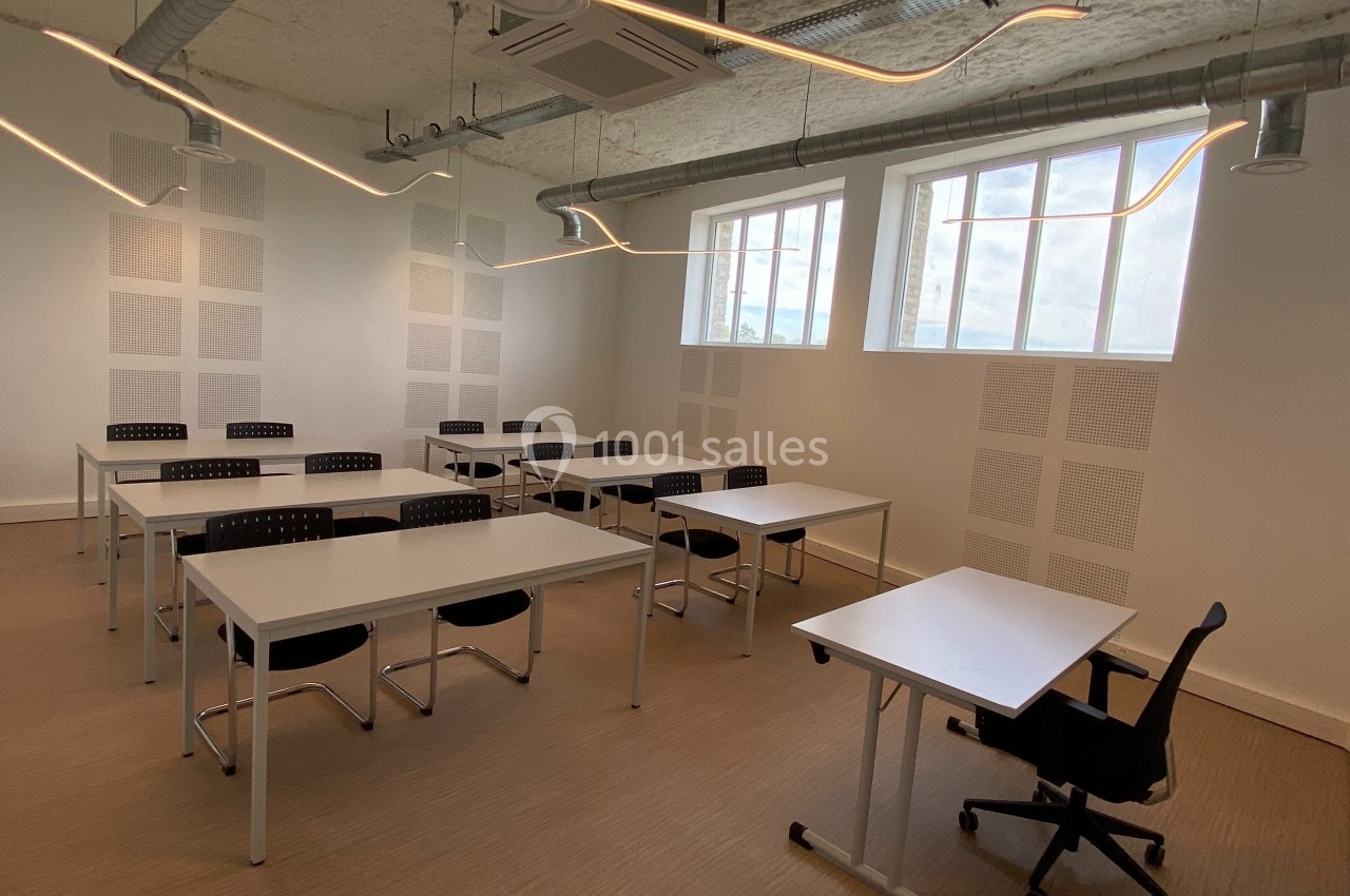 Salle de formation lumineuse avec des tables blanches, des chaises noires et un plafond équipé de luminaires modernes.