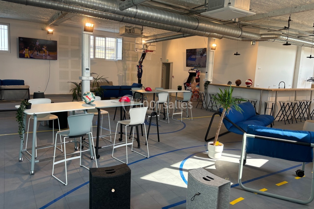 Espace intérieur lumineux avec tables hautes, chaises, canapés, plantes et décorations sportives.