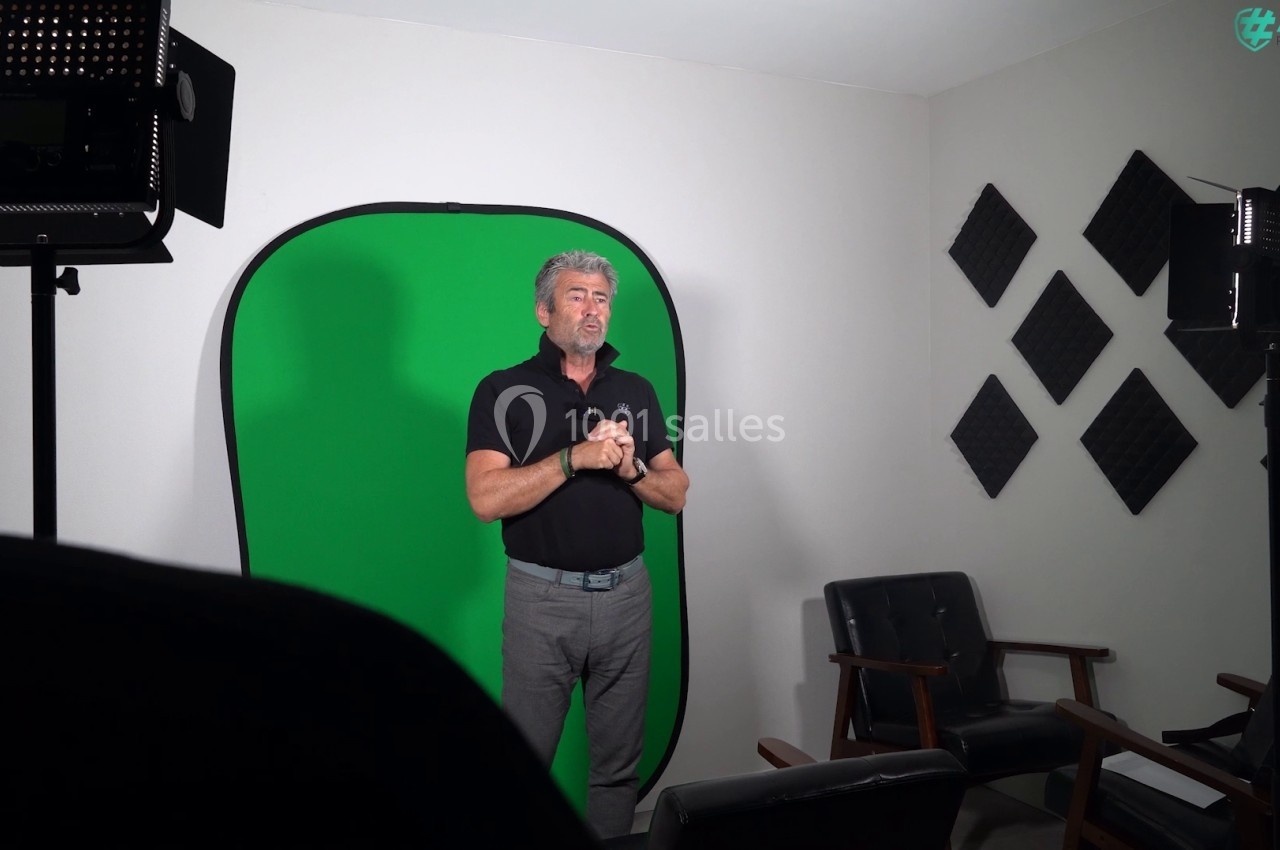 Un homme debout dans un studio avec un fond vert, éclairé par des projecteurs, entouré de panneaux acoustiques.