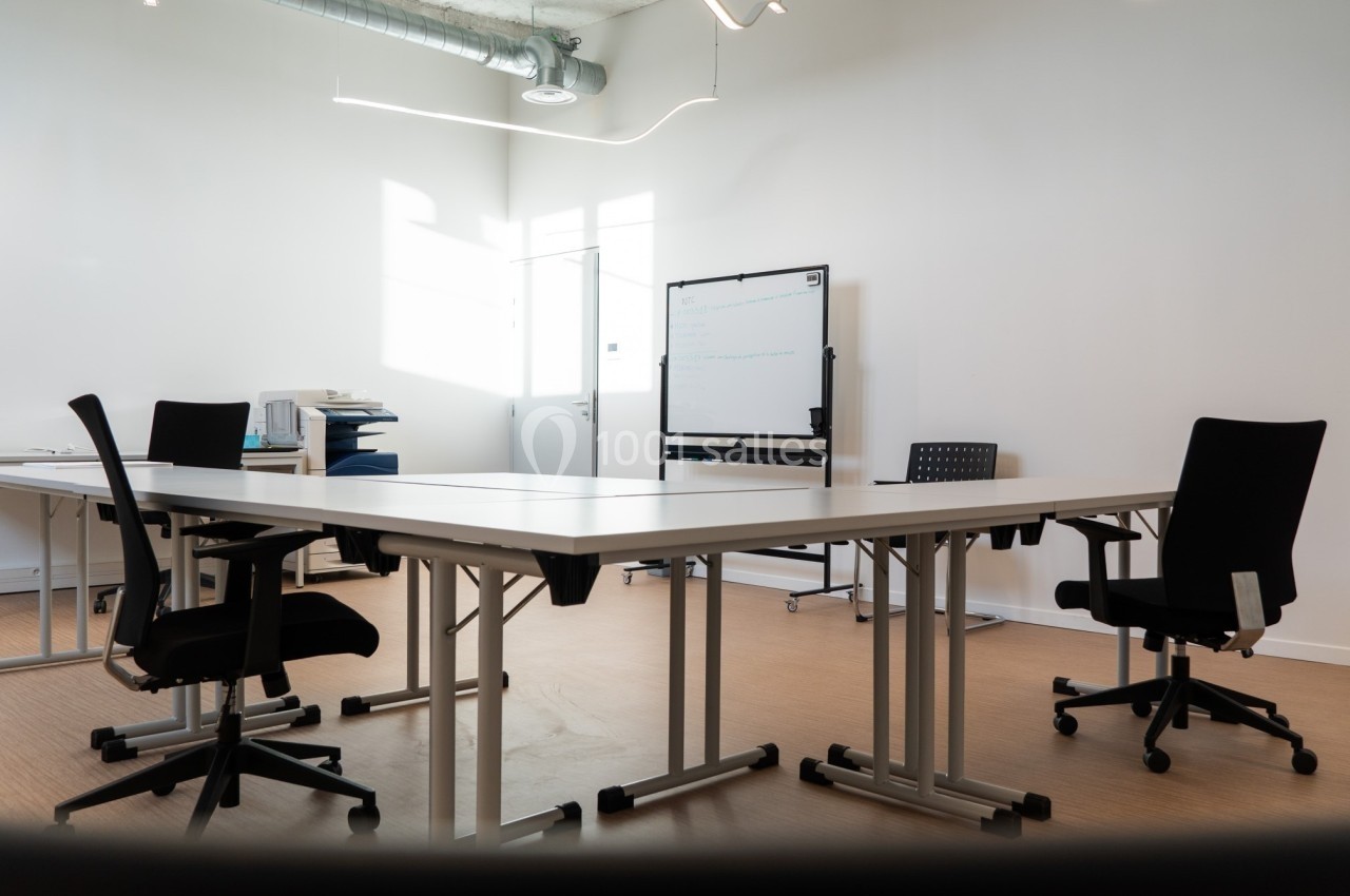 Salle de réunion lumineuse avec tables blanches, chaises noires, tableau blanc et équipement de bureau.