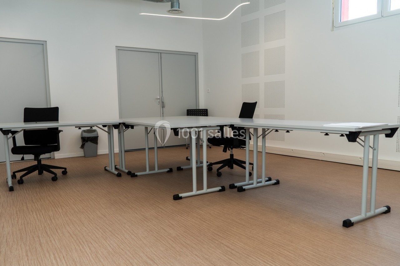 Salle de réunion avec tables modulables blanches, chaises noires et sol en parquet clair.