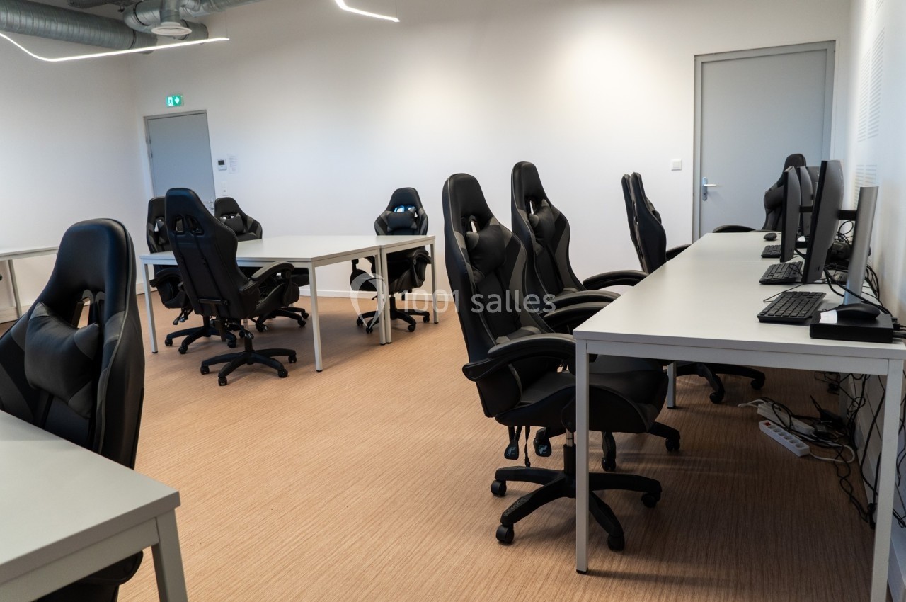 Salle équipée de bureaux, chaises ergonomiques et ordinateurs, éclairée par des néons, avec un sol en parquet clair.