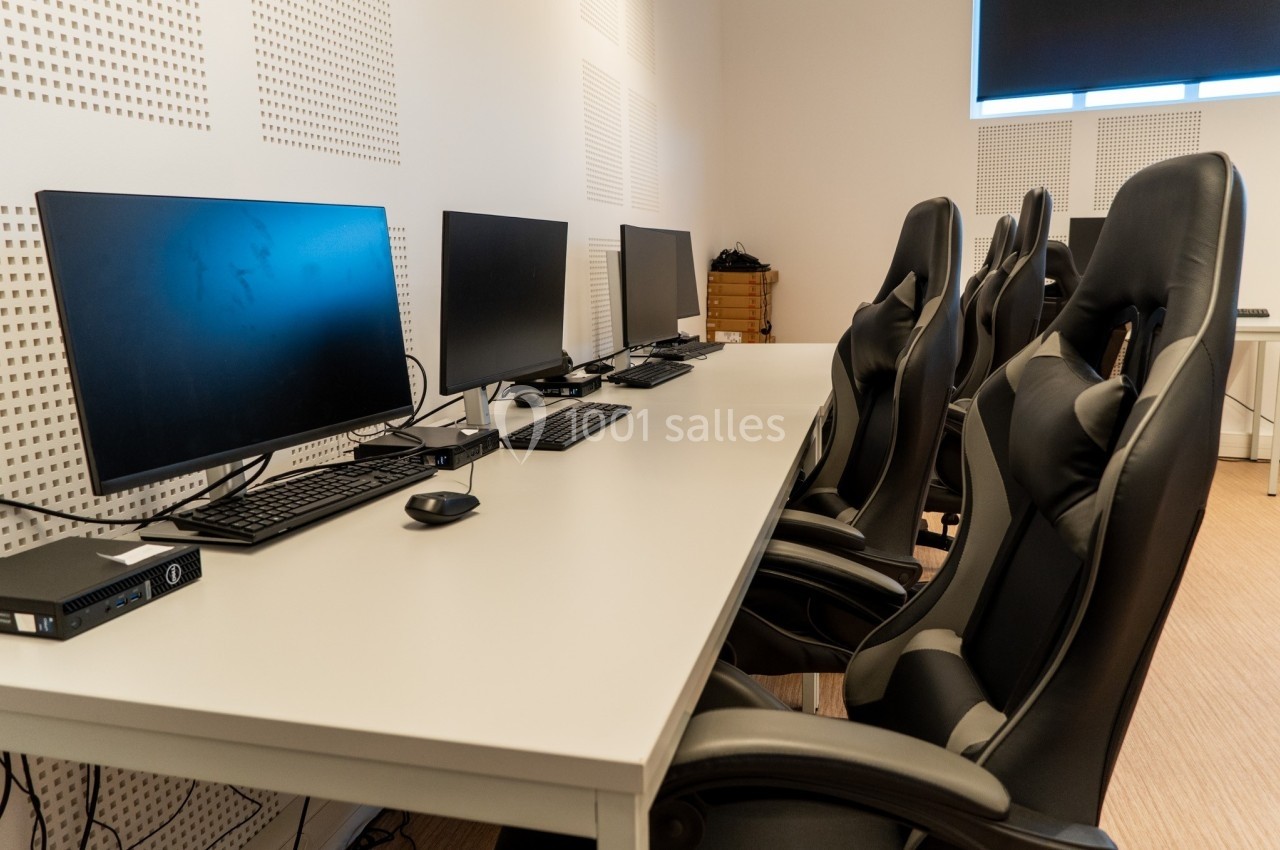 Salle équipée de bureaux alignés avec des ordinateurs, des écrans et des chaises ergonomiques noires.