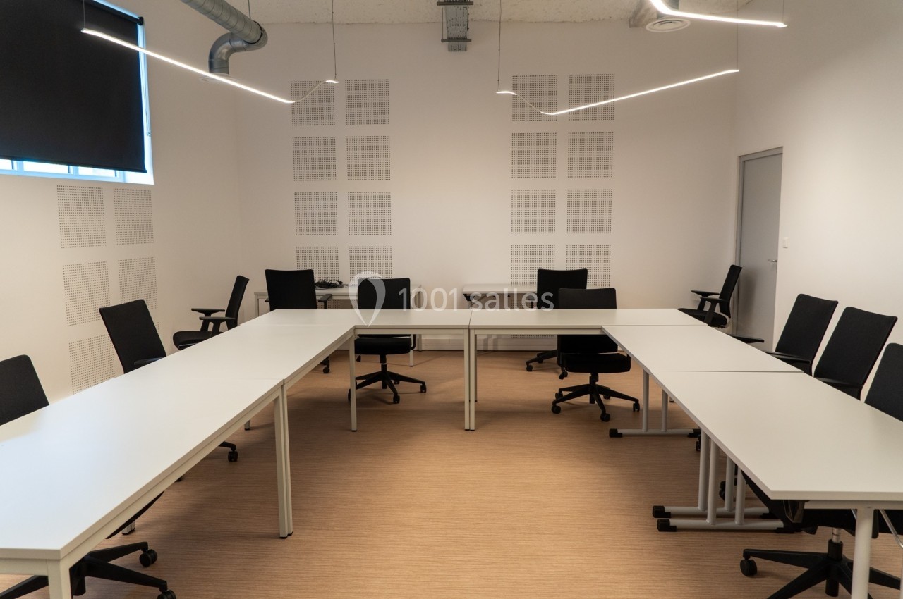 Salle de réunion lumineuse avec tables blanches disposées en U et chaises noires sur un sol en bois clair.