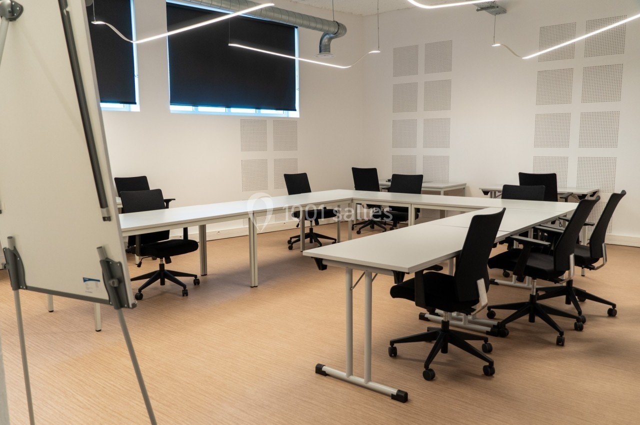 Salle de réunion moderne avec tables blanches disposées en U, chaises noires et tableau blanc au premier plan.