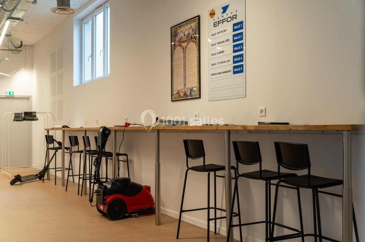 Salle équipée de tabourets hauts alignés le long d'un comptoir, avec un aspirateur et un tableau mural en arrière-plan.