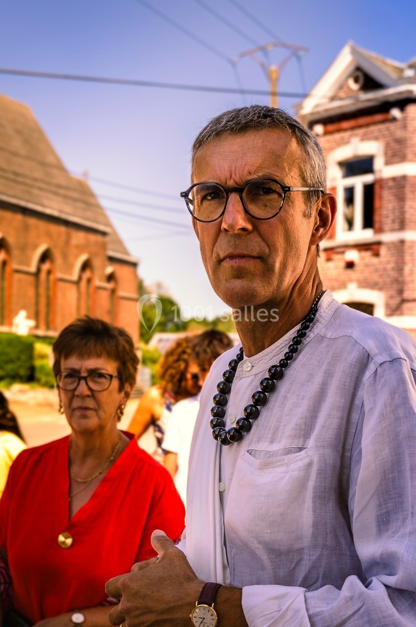 Un homme en chemise blanche et une femme en robe rouge se tiennent devant des bâtiments en briques sous un ciel bleu.