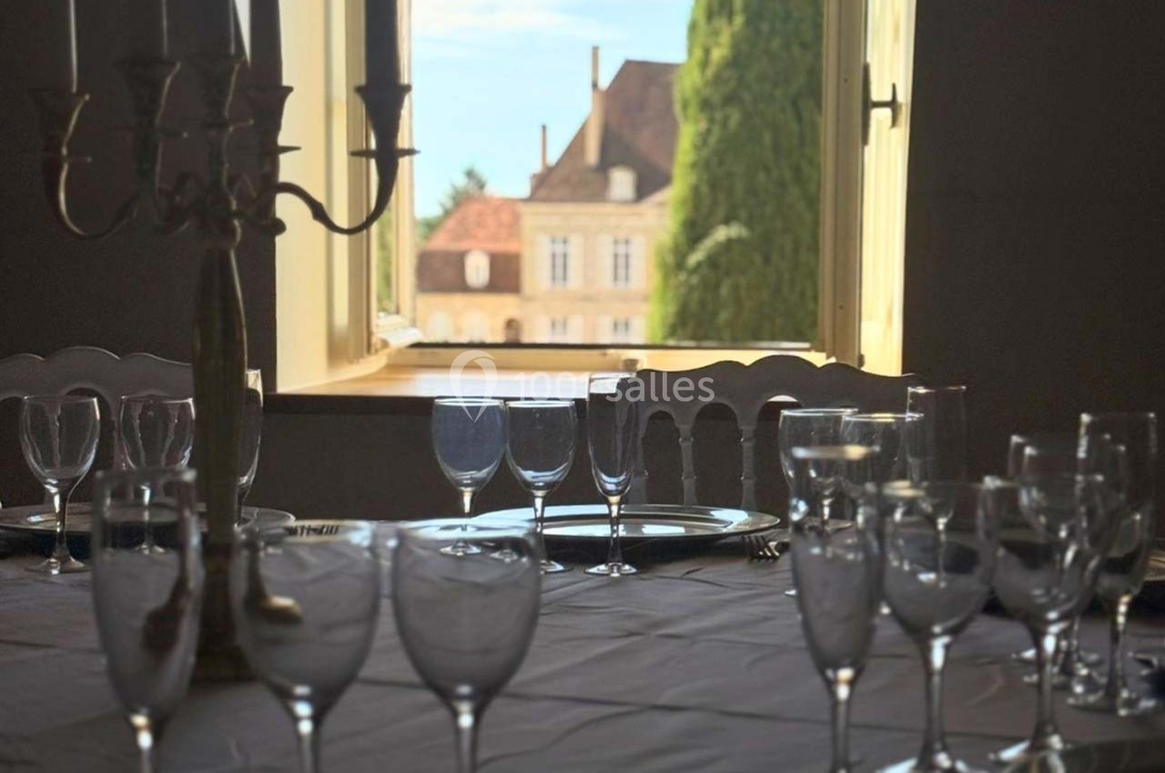 Table dressée avec des verres et un chandelier devant une fenêtre ouverte offrant une vue sur un bâtiment ancien et des…