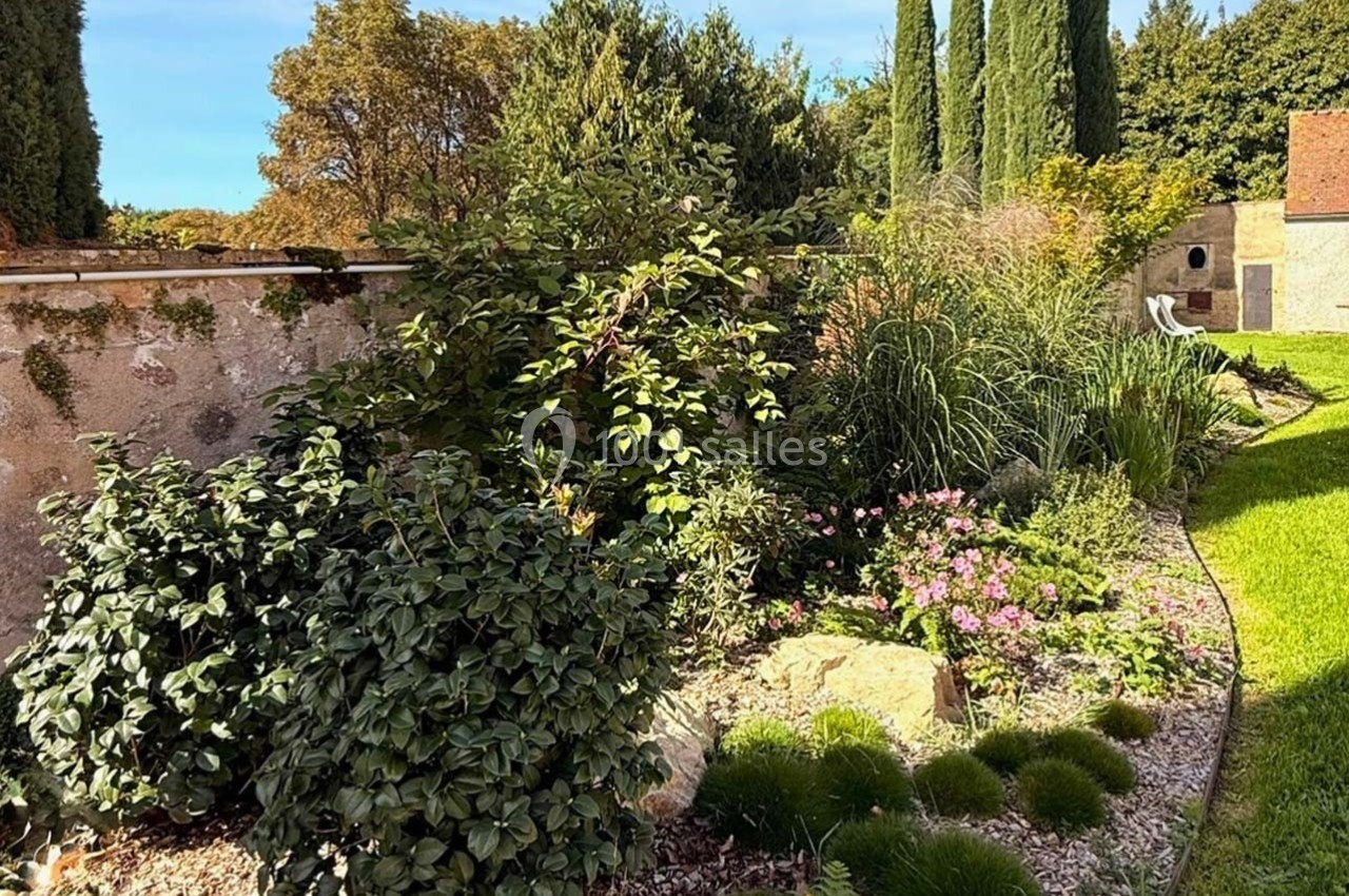 Jardin paysager avec arbustes, fleurs et pelouse, bordé par un mur en pierre et des arbres en arrière-plan.