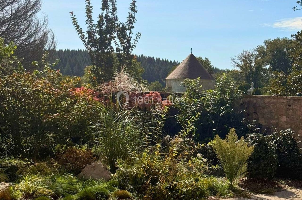Jardin paysager avec arbustes, fleurs et arbres, devant un mur en pierre et une tour au toit conique sous un ciel bleu.